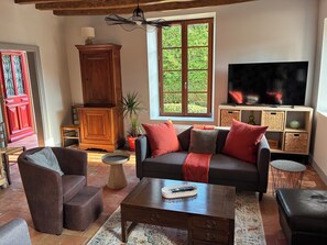 A cozy living room with a sofa, armchairs, a coffee table, a TV, and a window with a view of greenery.