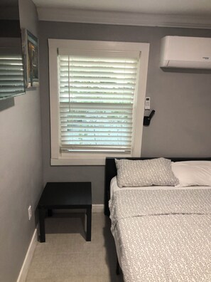 Un dormitorio con una cama, una ventana con persianas, una mesita de noche y un equipo de aire acondicionado.