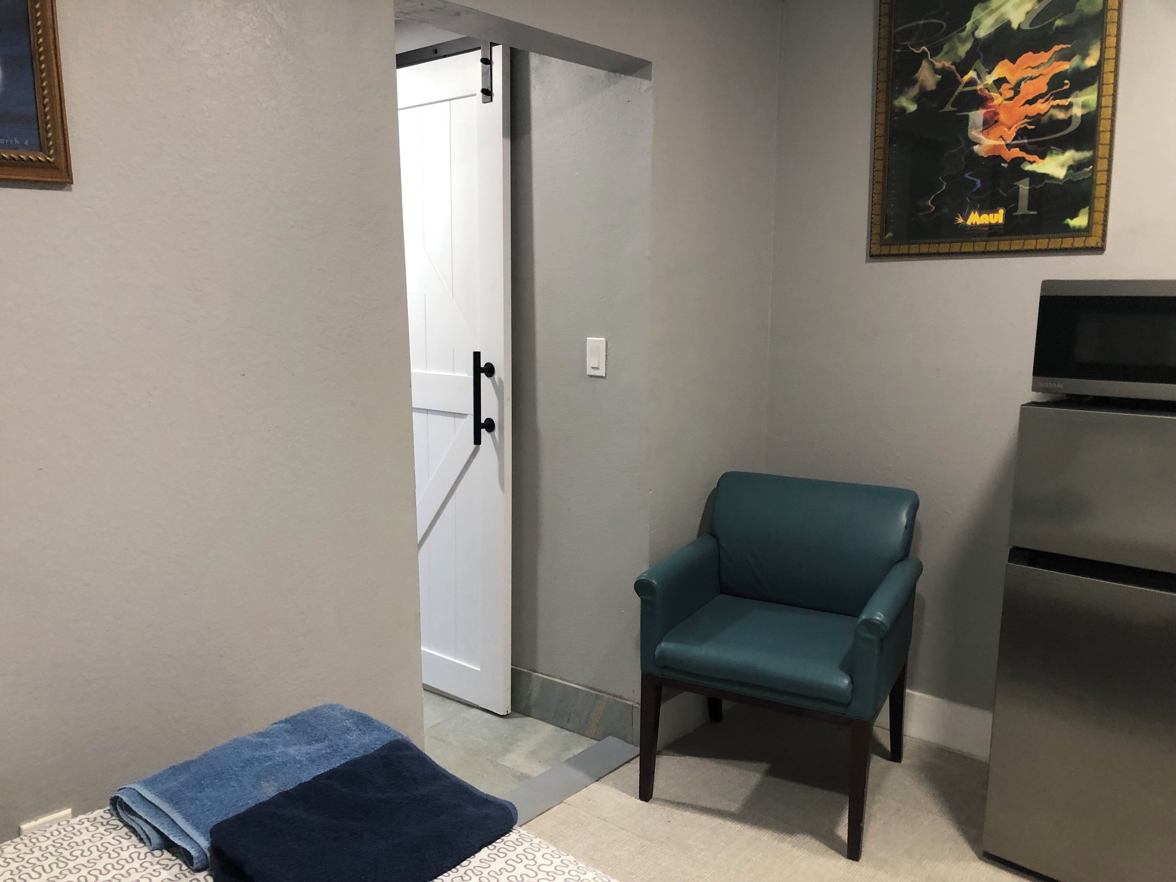 Une pièce avec un fauteuil turquoise, une porte blanche, un réfrigérateur et un tableau accroché au mur.