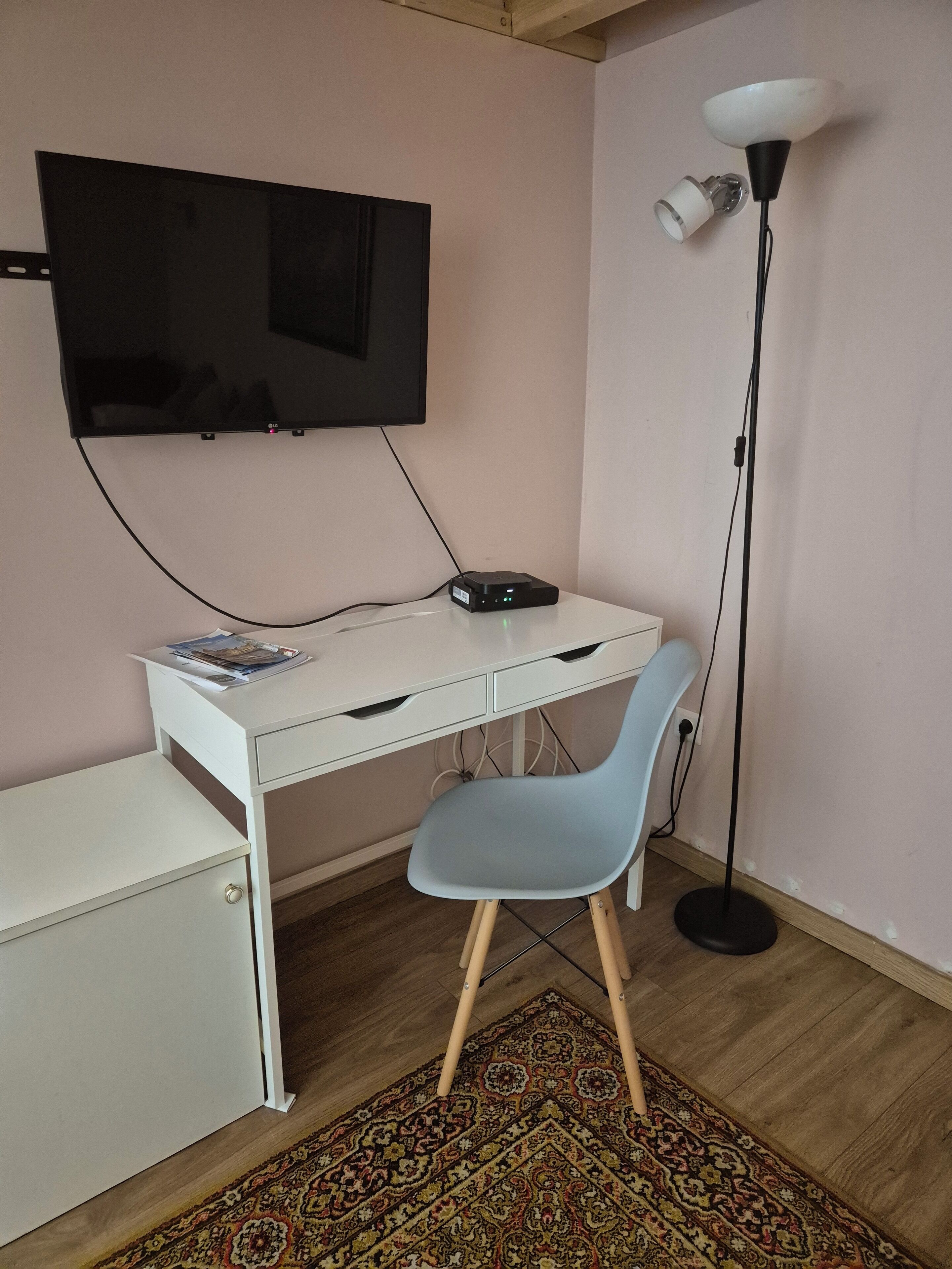 Une pièce avec un bureau blanc, une chaise bleue, un téléviseur à écran plat fixé au mur, un lampadaire et un tapis à motifs sur le sol.