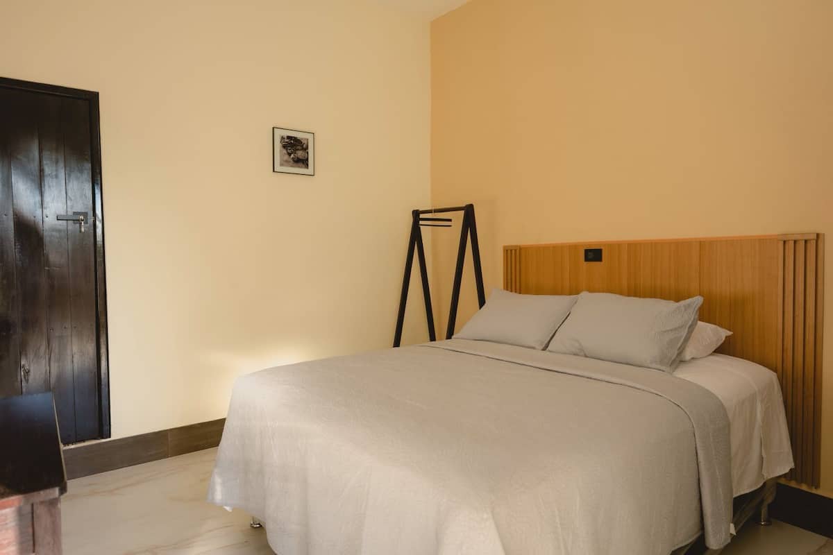 Ein ordentlich bezogenes Bett mit einem hölzernen Kopfteil, ein kleines gerahmtes Bild an der Wand und eine dunkle Holztür.