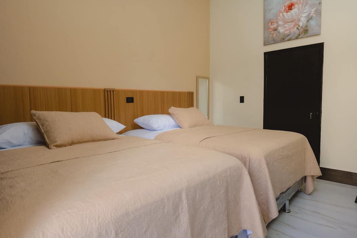 Ein Hotelzimmer mit zwei Betten, jeweils mit einer beigen Tagesdecke und weißen Kissen. Es gibt ein hölzernes Kopfteil, eine schwarze Tür und ein Blumenbild an der Wand.