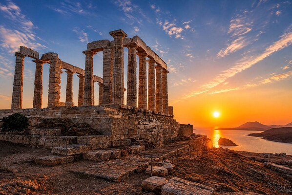 Des ruines antiques avec des colonnes, un sentier pavé de pierres et un coucher de soleil sur l’eau.