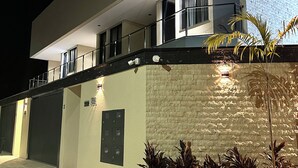 A modern building with a flat roof, large windows, and a balcony. There is a paved area with plants in front. - Apartamento Pequi Luxo com Garagem (Palmas)