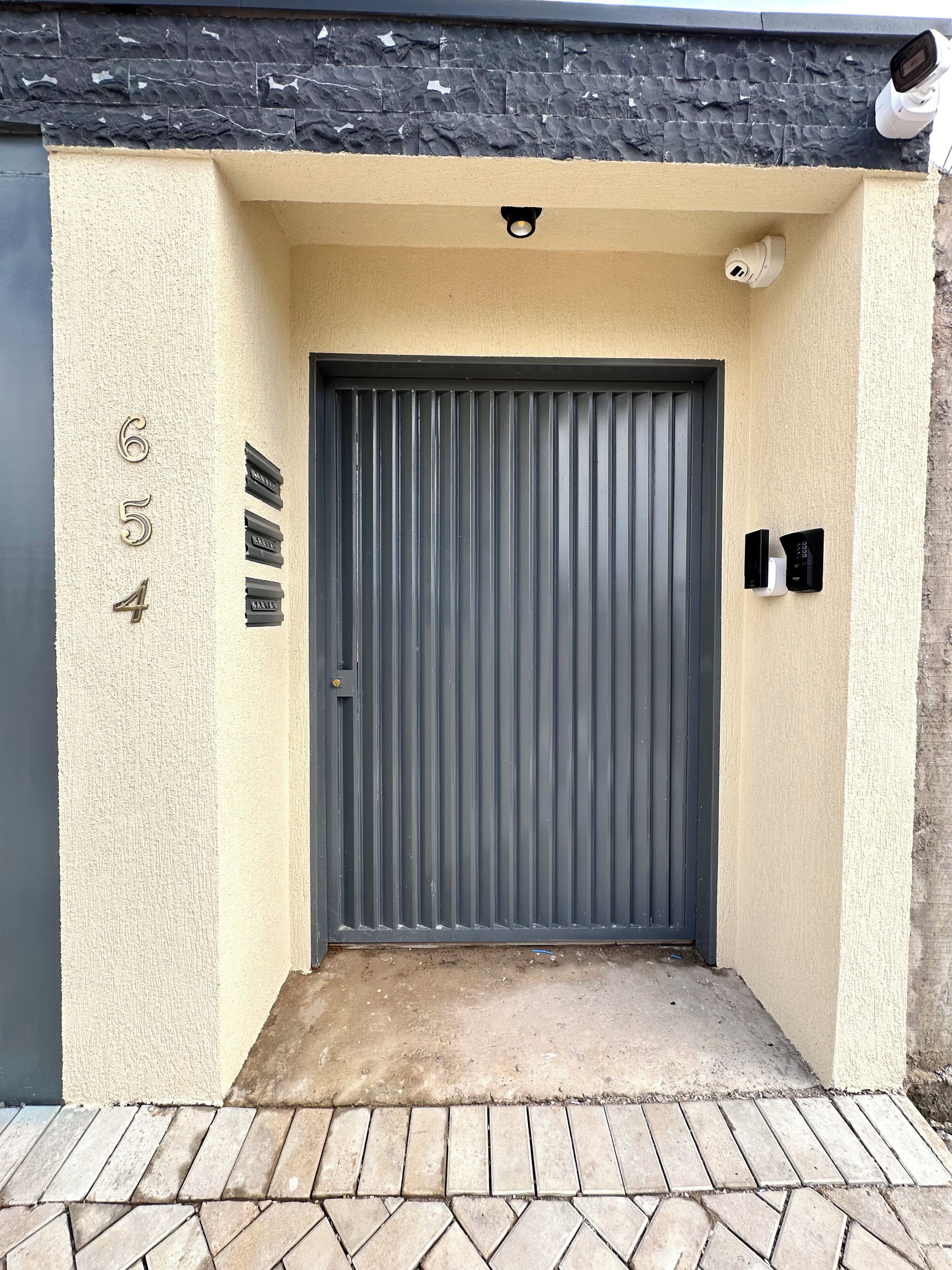 A grey metal door with a textured beige wall, a small security camera above, and a number '654' on the left.