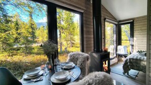 Et hyggeligt sommerhusinteriør med et spisebord dækket til to personer, en pejs og et stort vindue med udsigt over et skovlandskab.