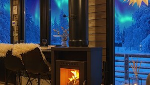 A cozy room with a wood-burning stove, a view of the aurora borealis through large windows, and a wooden deck outside. - Lapland Fell Village (Sodankyla)