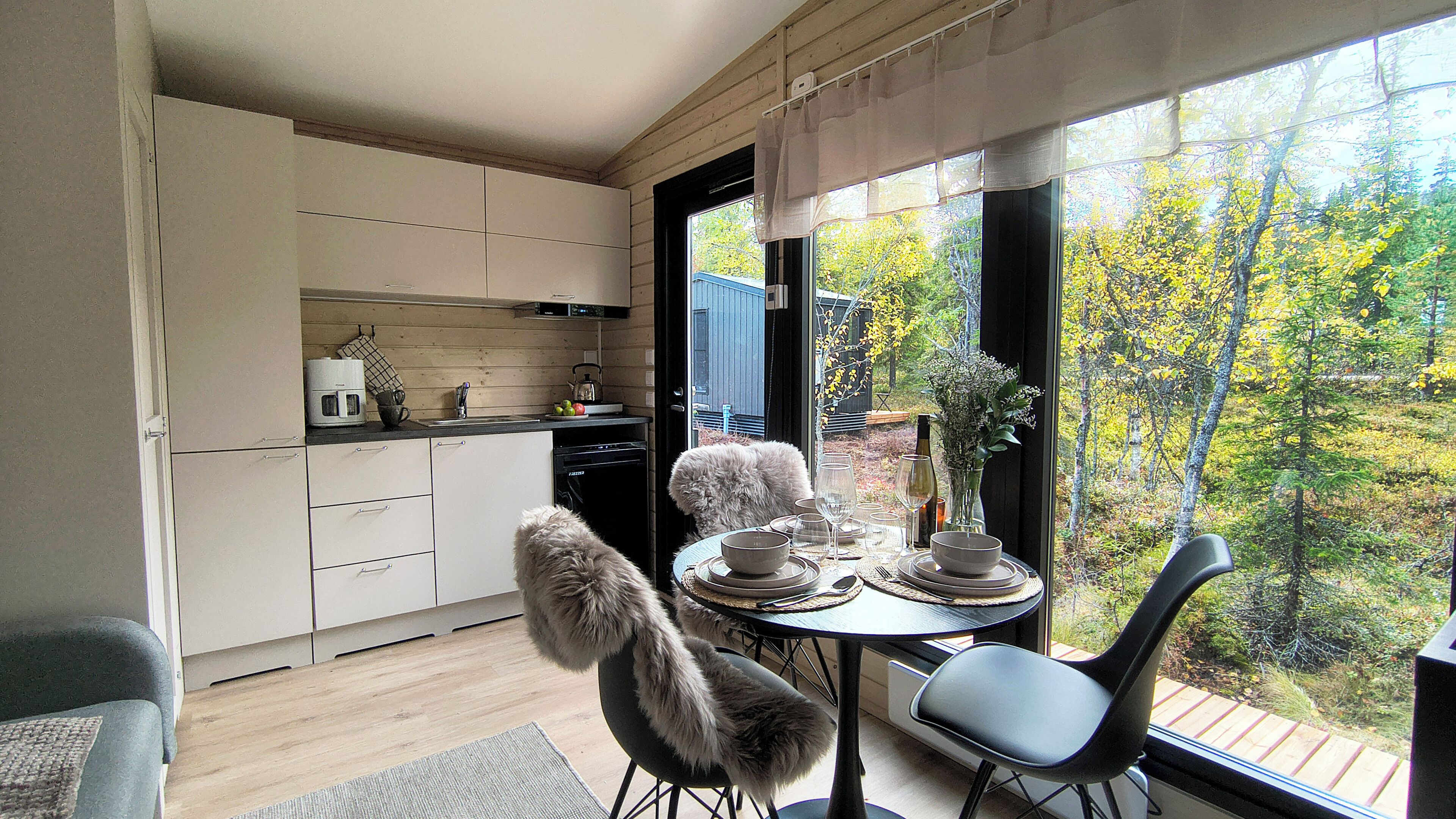 Una cocina moderna con armarios blancos, una mesa de comedor para dos personas y vistas a árboles otoñales a través de amplios ventanales.