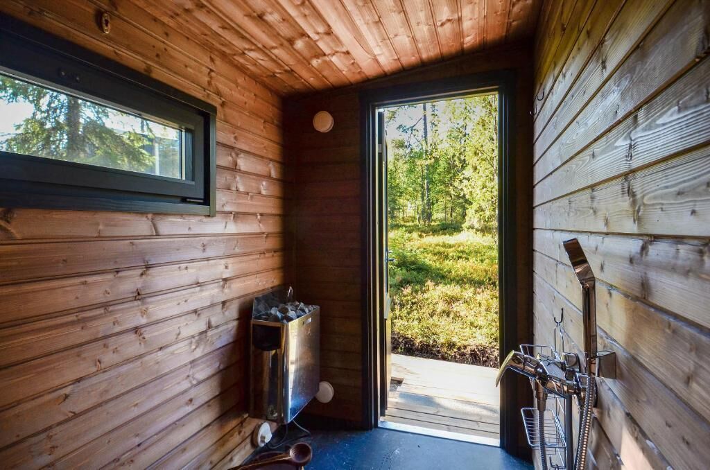 L’intérieur d’une cabane en bois, avec une grande fenêtre, un conteneur en métal et une porte donnant sur l’extérieur.