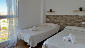 A hotel room with two single beds, a stone accent wall, a window with sheer curtains, and a view of the city.