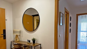 A hallway with a wooden door, a round mirror, a small table with a lamp, a potted plant, and a wicker basket.