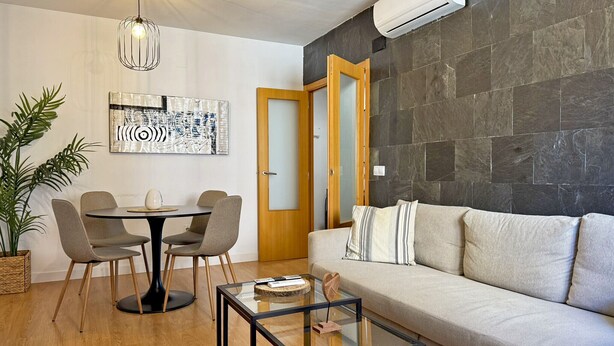 A modern living room with a round dining table, two chairs, a glass coffee table, a sofa, a potted plant, and a wall-mounted air conditioner.