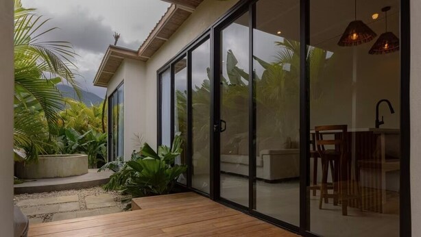 A modern house with large sliding glass doors, a wooden deck, and a garden with tropical plants. - CALALUN (La Fortuna)