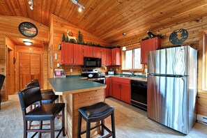 A cabin-style kitchen with wooden walls, a central island, red cabinets, stainless steel appliances, and a deer decoration on the ceiling. - Charming 2-bedroom cabin on a private Lake near Stone Lake. (Stone Lake)