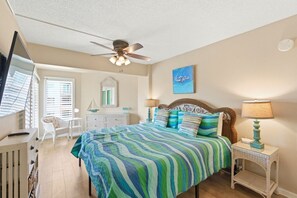 Ein Schlafzimmer mit Bett, Nachttisch, Stuhl, Kommode, Deckenventilator und Fenster mit Jalousien.