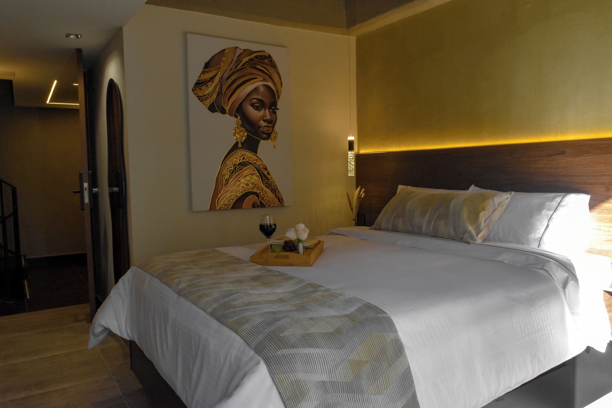 Ein Hotelzimmer mit einem großen Bett, einem hölzernen Kopfteil, einem Wandbild und einer Ablage mit Wein und Blumen.