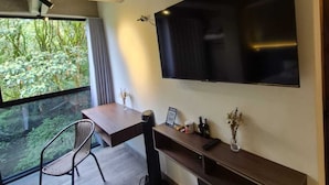 A modern room with a large TV mounted on the wall, a wooden desk, a chair, and a view of greenery outside. - Mali Experience (Rionegro)