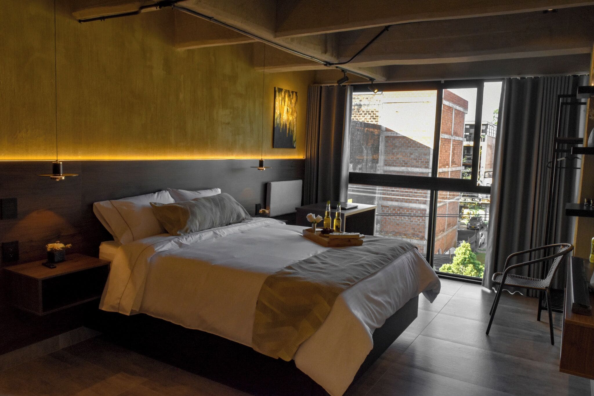 A modern bedroom with a large bed, bedside tables, a nightstand, a chair, and a view of the outside.