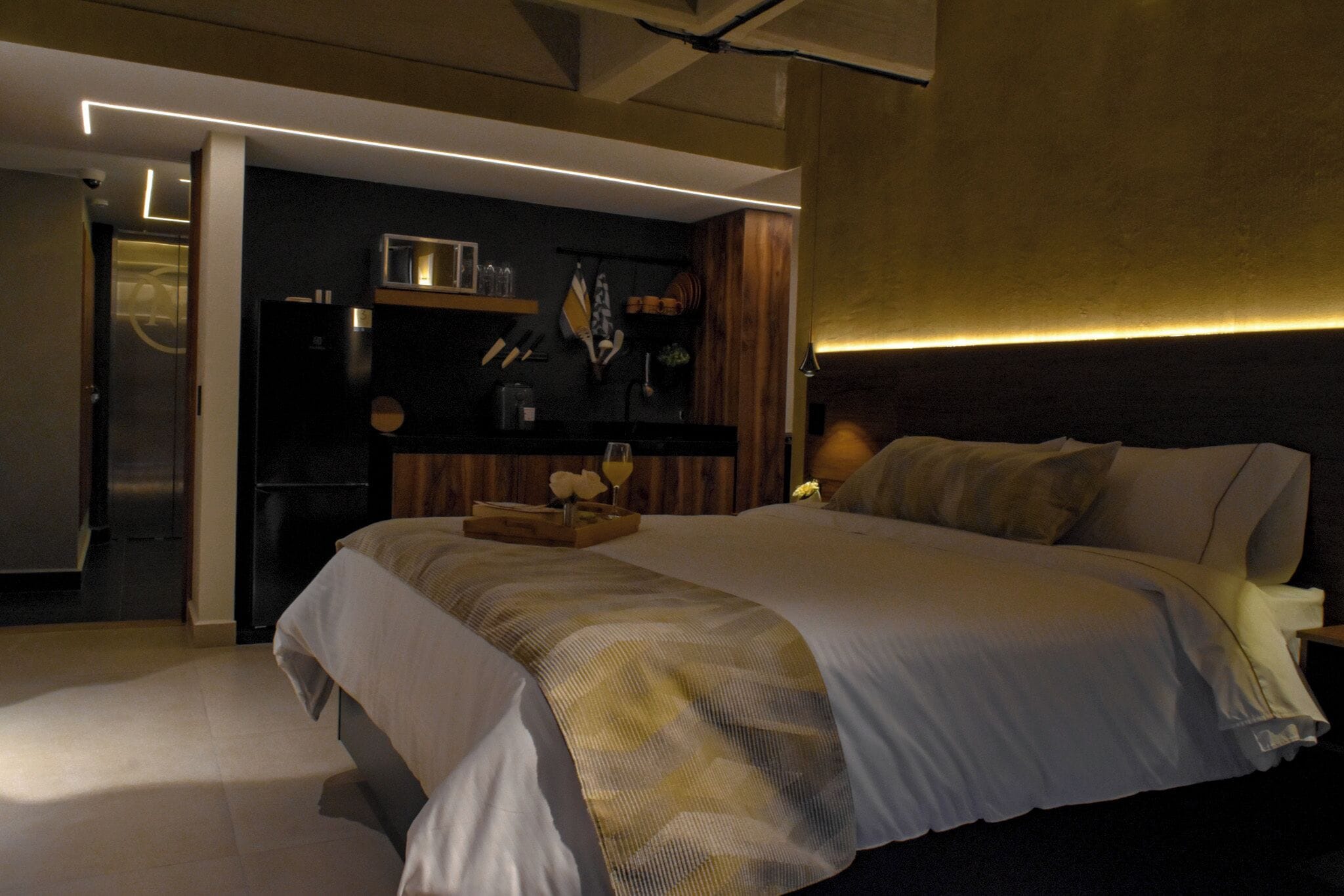 A modern hotel room with a large bed, a wooden headboard, and a built-in shelf with kitchenware.