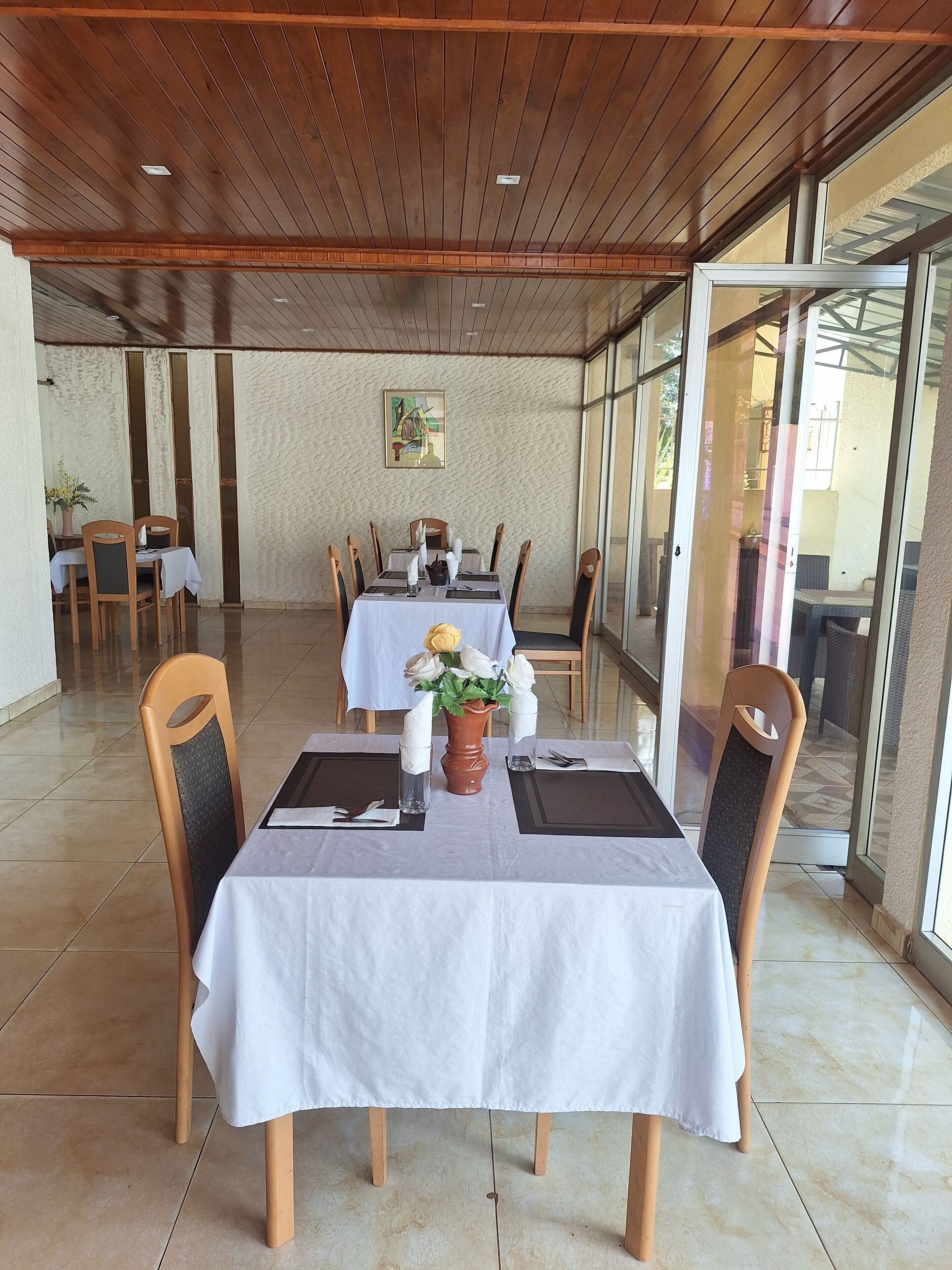 A dining area with tables set for two, featuring white tablecloths, wooden chairs, and a centerpiece with flowers.