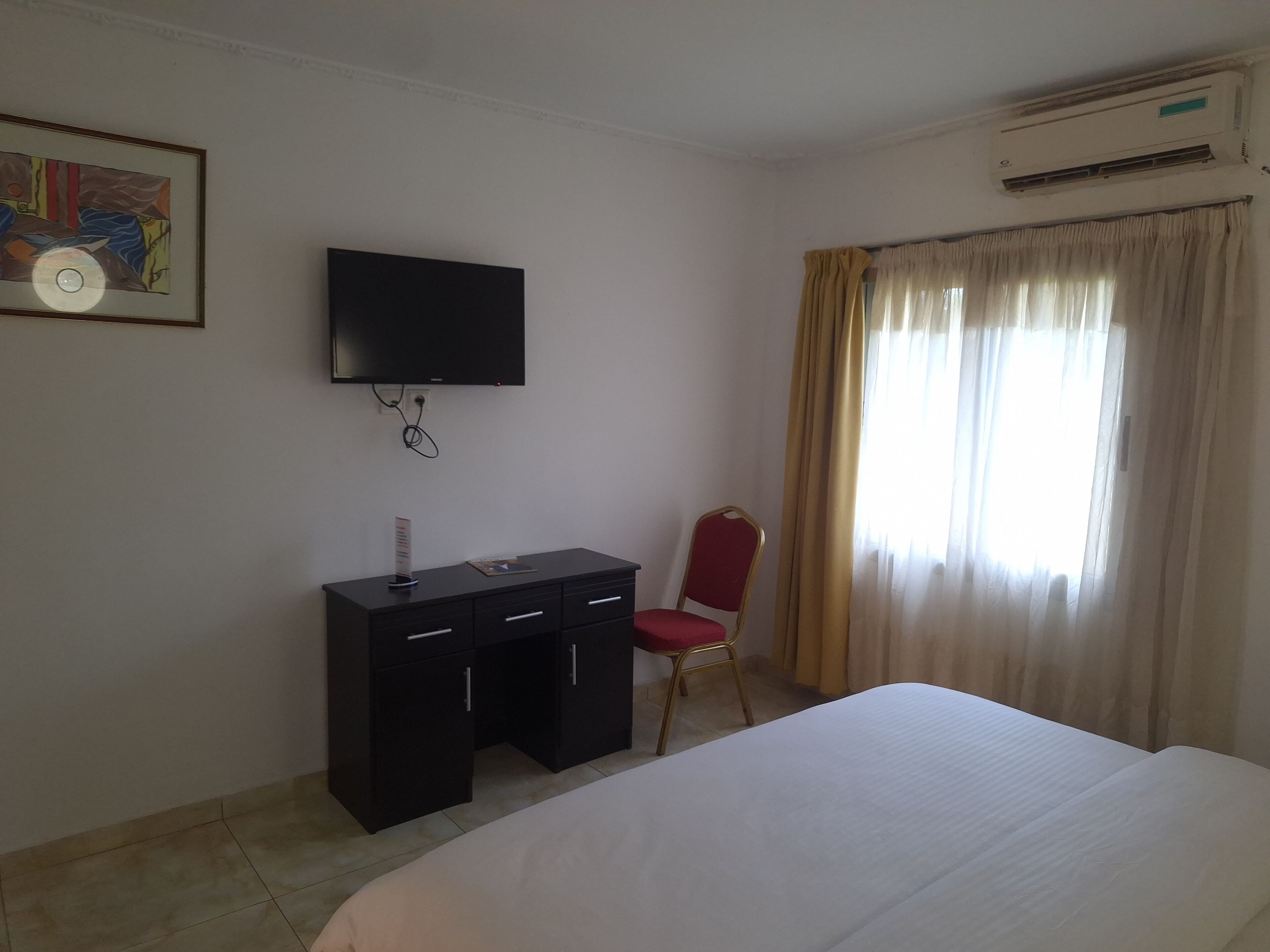 Habitación de hotel con cama, escritorio con cajones, silla, televisor montado en la pared y ventana con cortinas.