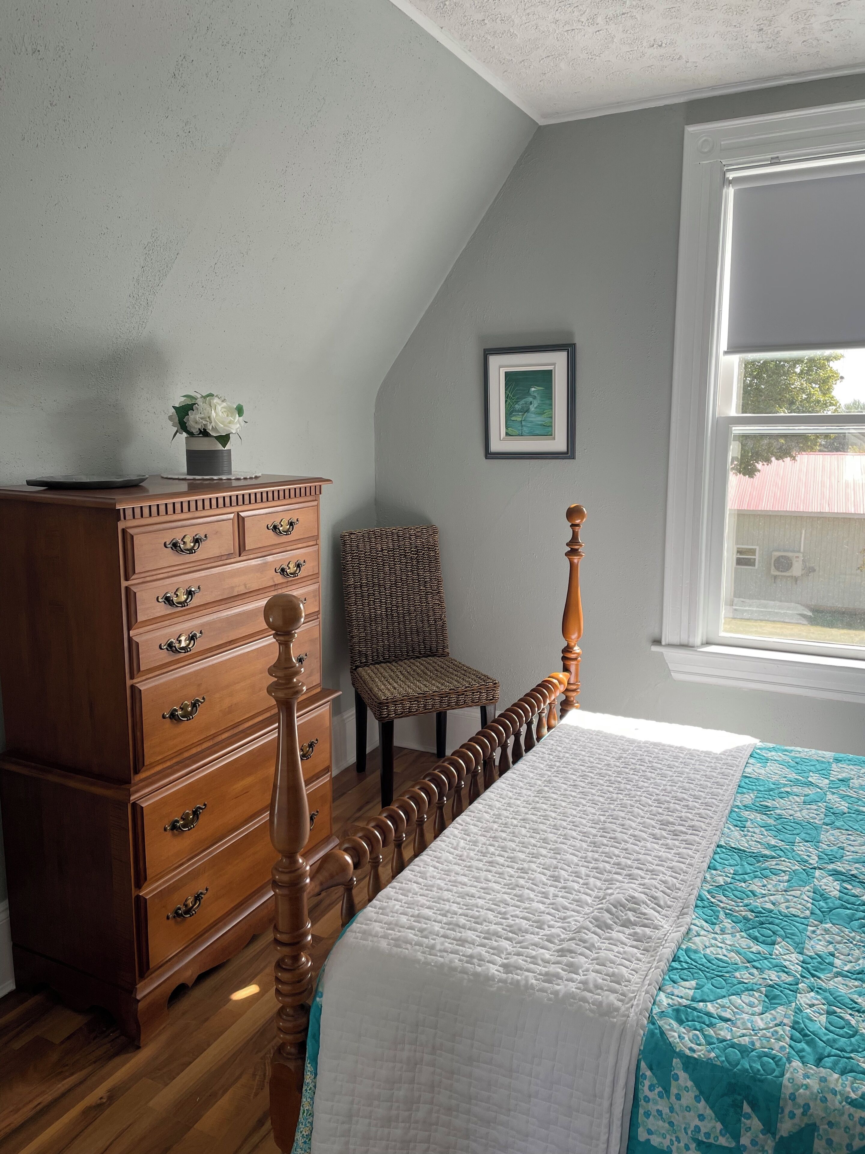 Ein Schlafzimmer mit einem Holzkoffer, einem Sessel, einem Bett mit Steppdecke, einem gerahmten Bild an der Wand und einem Fenster mit Blick auf ein Gebäude.