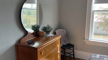 A bedroom with a wooden dresser, a round mirror, a potted plant, a chair, and a window with a view of greenery.