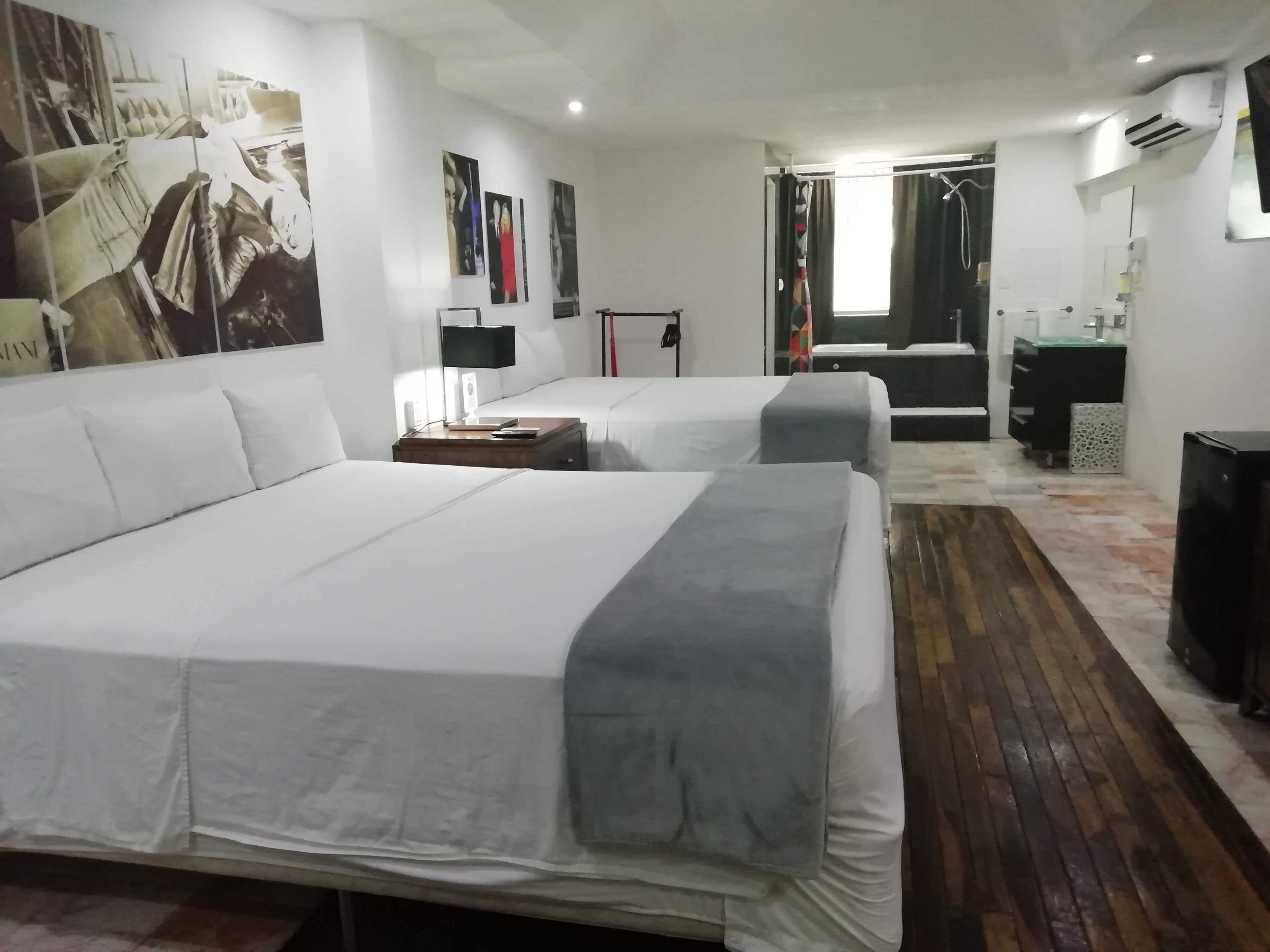 Una camera d'albergo con un letto grande, un letto più piccolo, una scrivania con una lampada, un armadio e un bagno visibile attraverso una porta aperta.