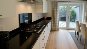 Cuina moderna amb fogó negre, armaris blancs i campana extractora d'acer inoxidable. Es veu una zona de menjador amb taula i caires a través d'una porta de vidre.