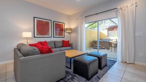 Ein modernes Wohnzimmer mit einem grauen Sofa, einem Holztisch und schwarzen Hocker. An der Wand hängen zwei gerahmte Bilder, eine Stehlampe und eine Glastür, die zu einem Außenbereich führt.