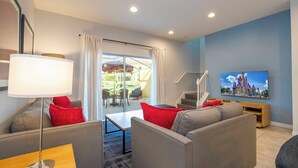 Ein modernes Wohnzimmer mit einer Couch, einem Couchtisch und einem Fernseher. Eine Treppe führt auf eine obere Ebene, und eine Glastür öffnet sich zu einem Balkon mit Gartenmöbeln.