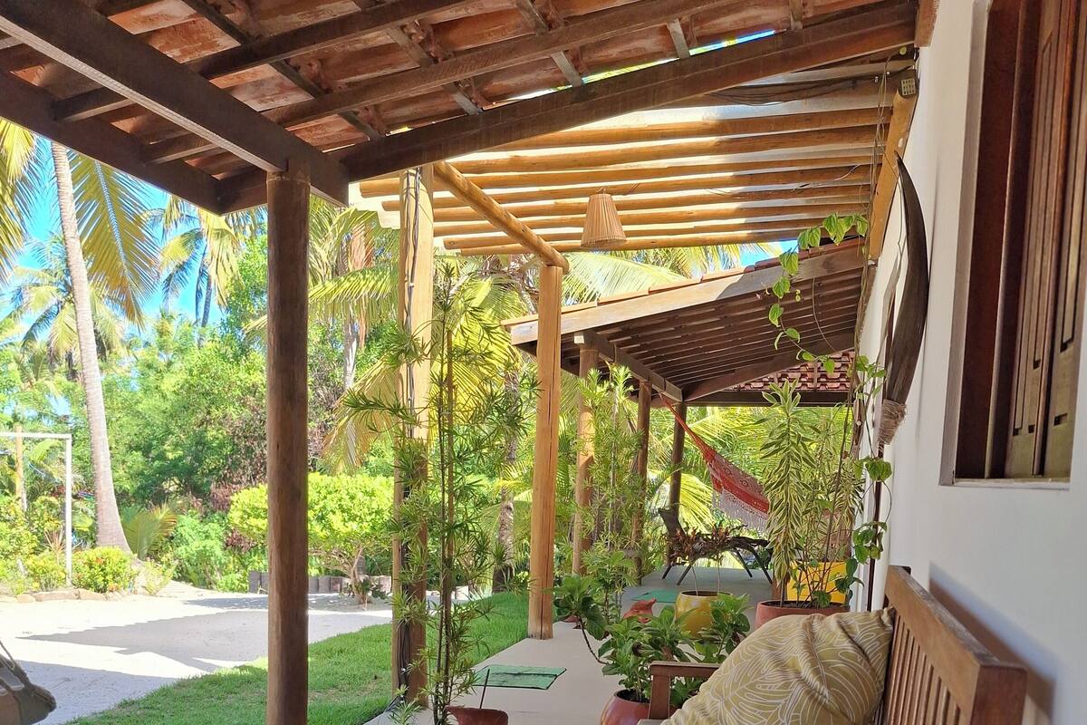 A covered outdoor seating area with a wooden bench, potted plants, and a view of a lush garden.