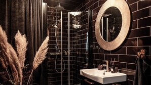 A modern bathroom with a black tiled wall, a round mirror, a white sink, and a glass shower enclosure. - Falknästet unique accommodation in one of Sweden's most spectacular natural areas. (Höganäs)