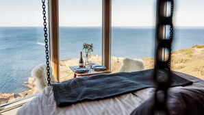 A bedroom with a large window overlooking the sea, a bed, a table with a bottle and glasses, and a fluffy chair. - Falknästet unique accommodation in one of Sweden's most spectacular natural areas. (Höganäs)