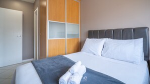 A neatly made bed with white linens and a grey blanket, a grey headboard, and a wooden wardrobe with glass doors. - Apts in a Building 100 Meters From Ufsc - Eoss (Florianópolis)