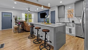 A modern kitchen with a central island, bar stools, a dining area with a sofa, and a TV mounted on the wall.