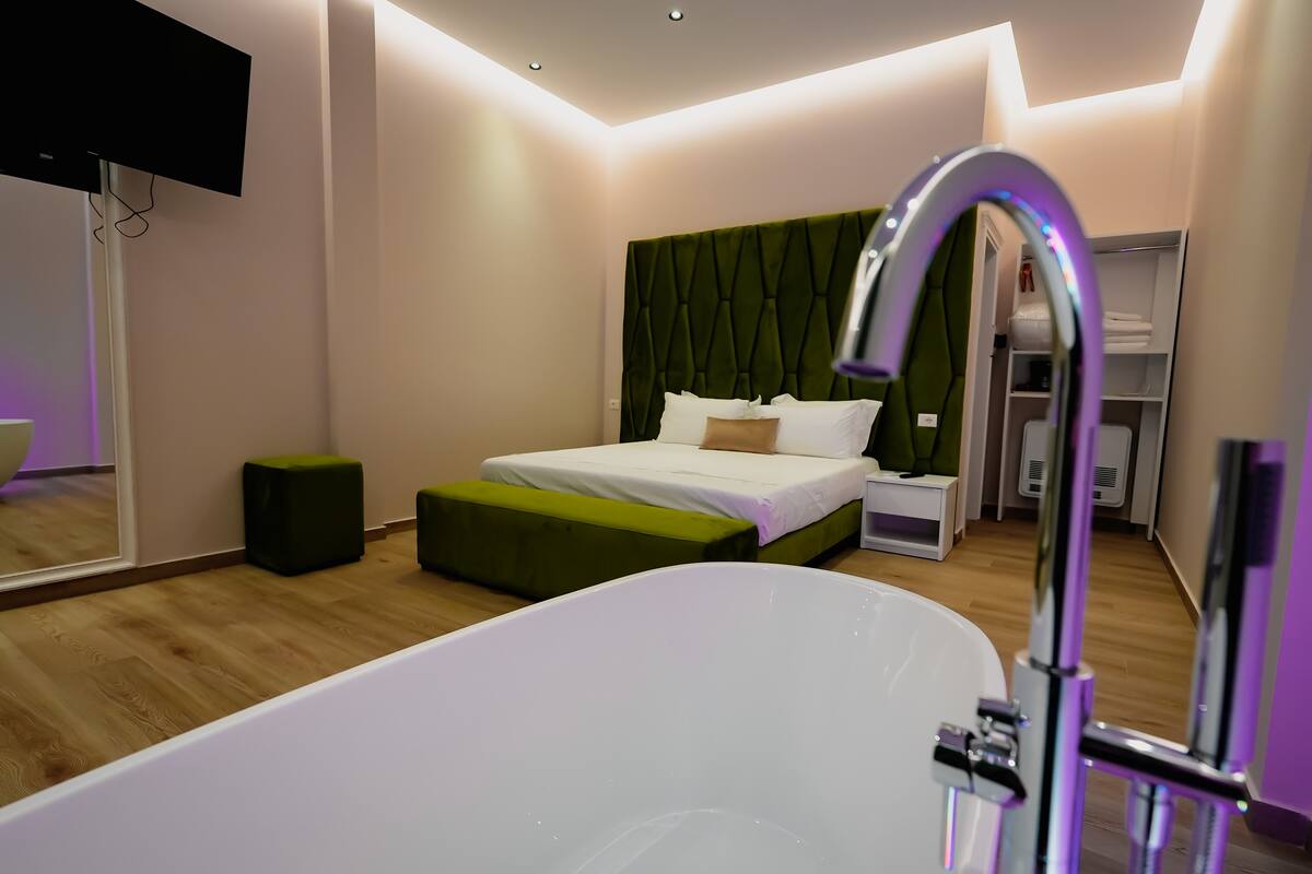 Una camera da letto moderna con un letto grande, una panca imbottita verde, una vasca da bagno bianca e una toeletta con specchio.