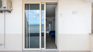 A room with a sliding glass door leading to a balcony, a kitchenette, and a bed with a blue cover. - Aria Condizionata e Terrazza Vista Mare (Castelsardo)