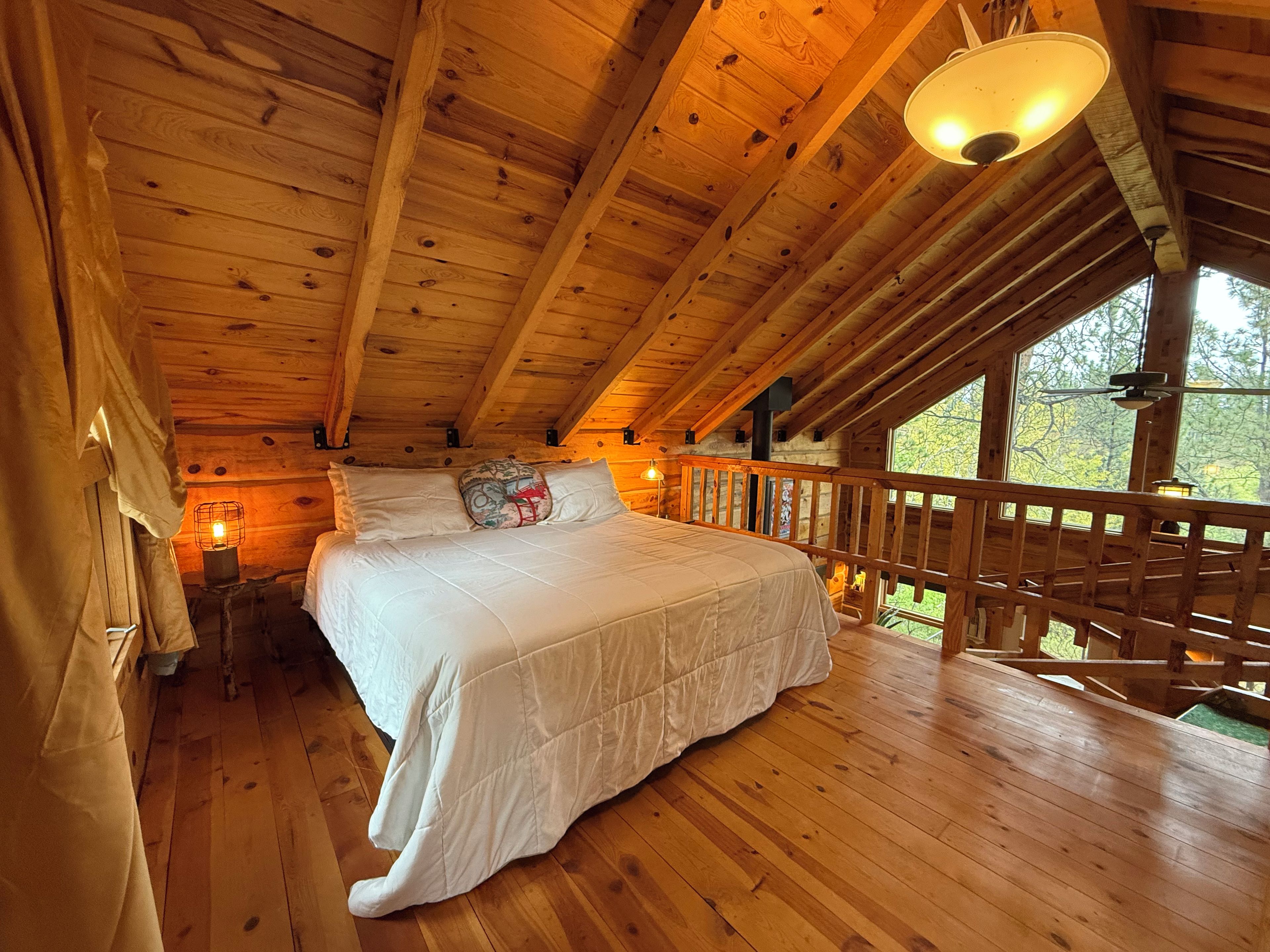 A wooden cabin bedroom with a bed, a nightstand, a window, and a balcony railing.