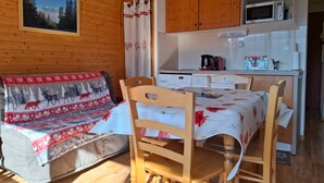 A cozy room with a wooden wall, a sofa with a festive blanket, a dining table with chairs, a kitchen area with cabinets and appliances, and a window with a view of trees. - V du Bachat Epicea B Ndeg22 - 3P6 (Chamrousse)