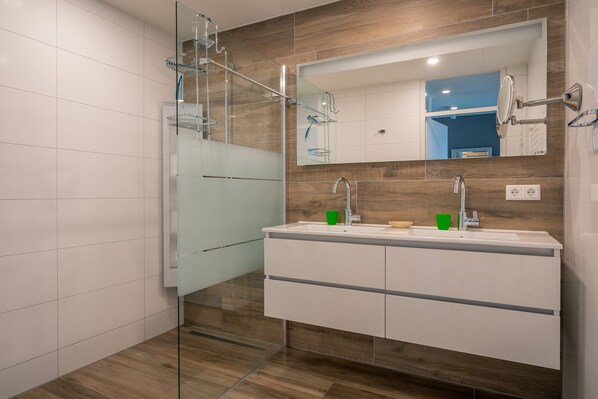Une salle de bain moderne dotée d’une douche à parois de verre, d’un grand miroir et d’un meuble-lavabo blanc avec un pare-éclaboussures en bois.