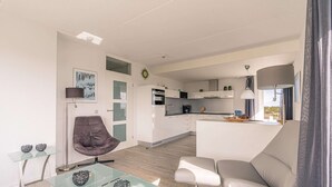 Uma sala de estar moderna com uma mesa de centro de vidro, uma poltrona cinza e um sofá. Uma cozinha com armários brancos e uma área de jantar com mesa e cadeiras são visíveis ao fundo.