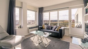 Sala de estar moderna com uma mesa de centro de vidro, sofá com almofadas e amplas janelas com vista para a cidade.