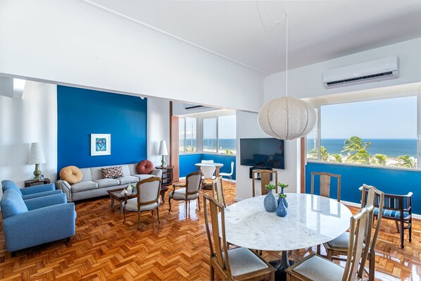 A spacious living room with a wooden herringbone floor, a blue accent wall, a large window with a view of the ocean, a dining area with a marble table, and a modern air conditioning unit. - Luxury 3bdrs Sea View in Copacabana A 2440/515 (Rio de Janeiro)