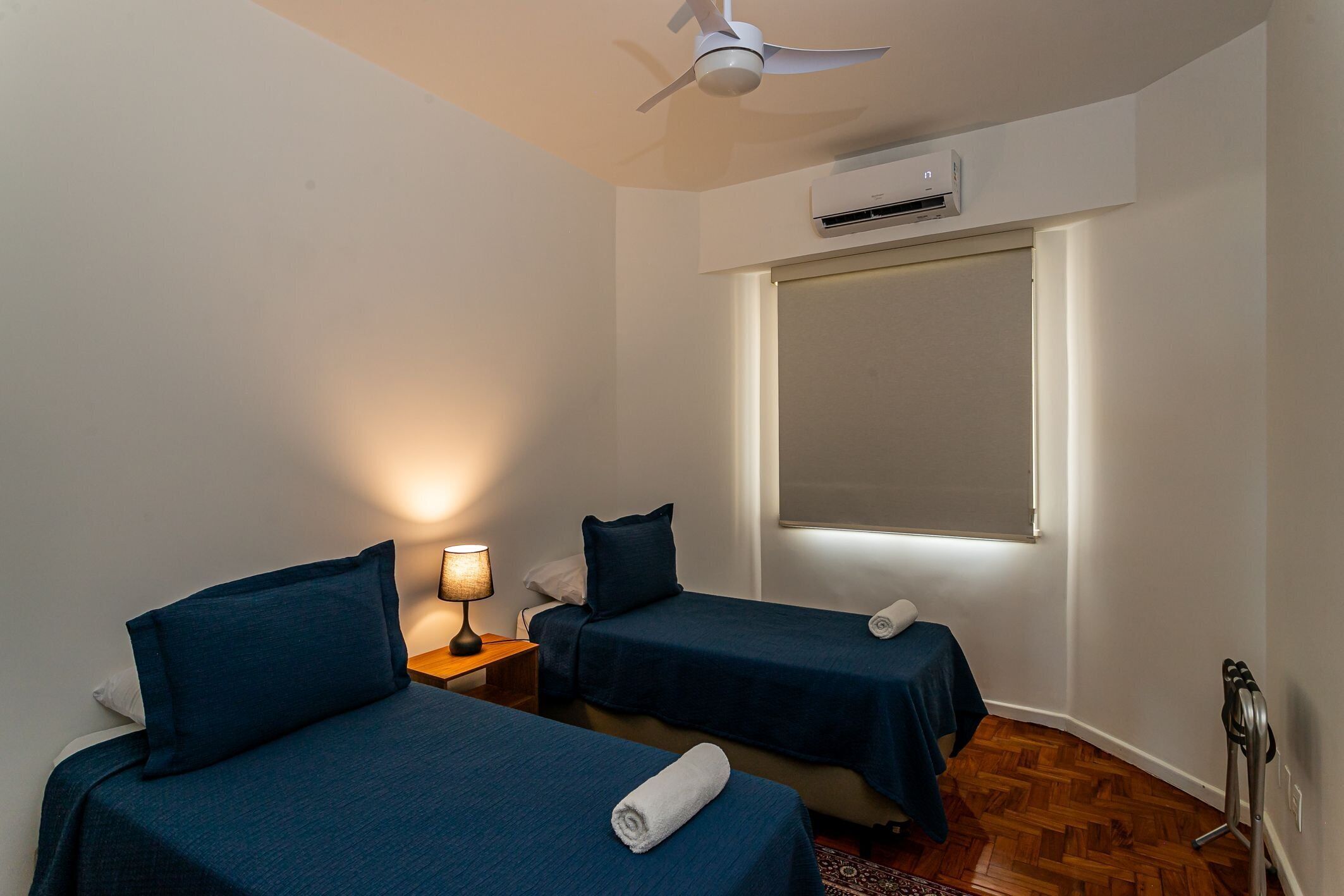 Una habitación de hotel pequeña con dos camas individuales, un ventilador de techo, un equipo de aire acondicionado y una ventana con persianas.