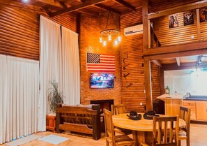 Un interior rústico con una mesa de comedor de madera, sillas, un banco, una bandera estadounidense, un televisor y una zona de cocina.