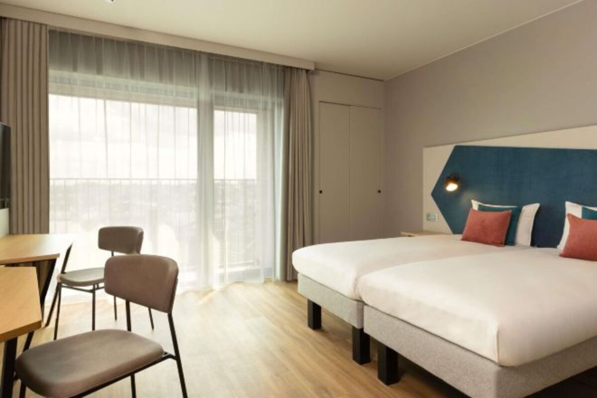 Une chambre d’hôtel avec deux lits, un bureau, une chaise et une grande fenêtre avec des rideaux.