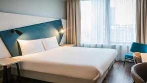 A hotel room with a large bed, a bedside table, a chair, and a view of the city through large windows. - Aparthotel Adagio Antwerp City Center (Antwerp)