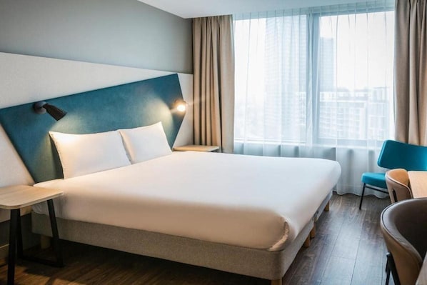 Habitación de hotel con una cama grande, una mesita de noche, una silla y vistas a la ciudad a través de amplias ventanas.