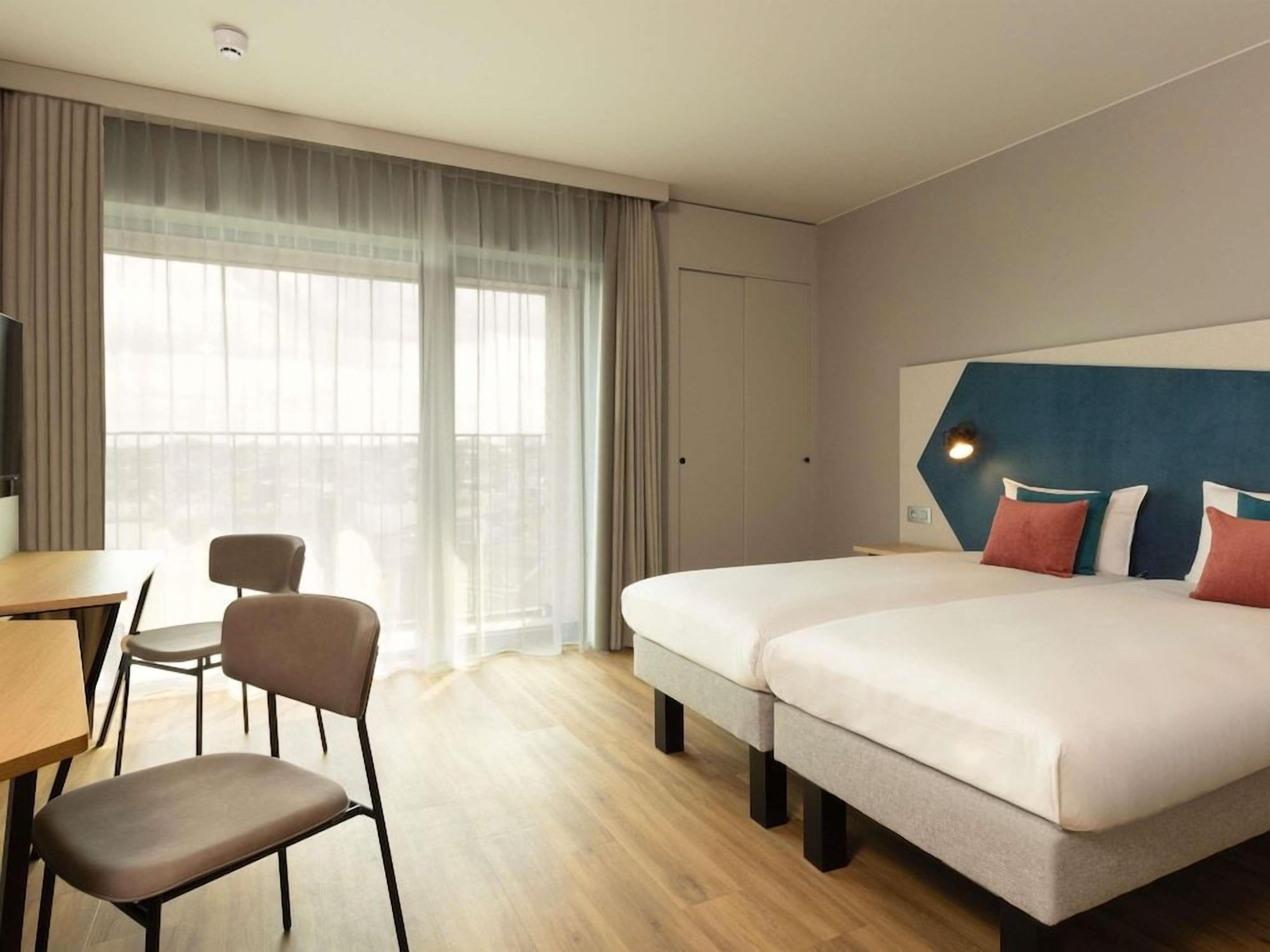 A hotel room with two beds, a desk, a chair, and a large window with curtains.